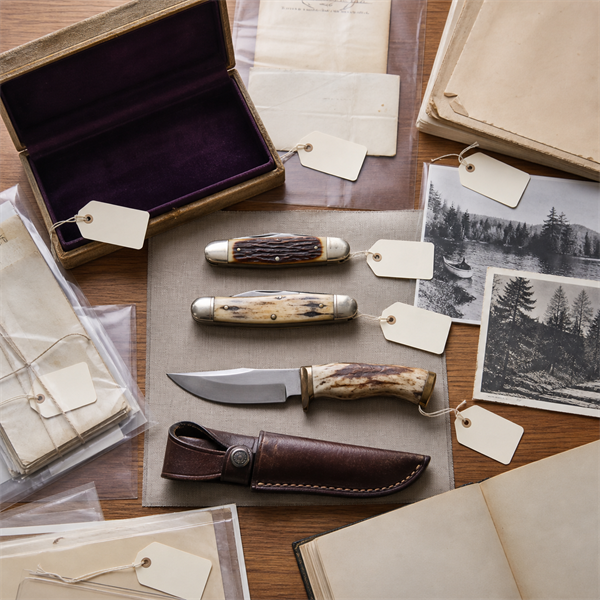 Unbranded collectible knives arranged with archival tags, old photographs, receipts, a presentation box, and a separate sheath.